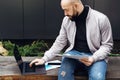 Bearded businessman freelancer working on laptop outdoors. Man holds paper documents in his hand and typing on laptop keyboard