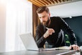Bearded businessman is standing by computer, looking thoughtfully at laptop screen, holding notebook, watching webinar. Royalty Free Stock Photo