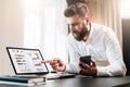 Bearded businessman in white shirt is sitting at table in front of computer, showing finger on laptop screen with graphs Royalty Free Stock Photo