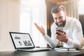 Bearded businessman in a white shirt is sitting at a table in front of a laptop with graphs, charts, diagrams on screen Royalty Free Stock Photo