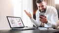 Bearded businessman in a white shirt is sitting at a table in front of a laptop with graphs, charts, diagrams on screen Royalty Free Stock Photo