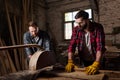 bearded carpenter in protective googles and gloves using machine saw and partner standing