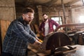 bearded carpenter in safety googles using machine saw and partner with crossed arms standing behind