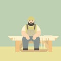 Bearded carpenter sitting on bench with saw and wood planks. Craftsman in apron resting at workbench in workshop