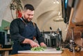 Bearded chef cook dressed black uniform with an apron chopiing fresh green cucumber with sharp knife in basement restaurant