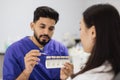 Bearded confident male dentist checking and selecting color of young woman teeth