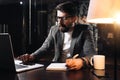 Bearded coworker sits by the wooden table with lamp and using contemporary notebook. Businessman hold pen and analysing trends