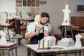 Bearded craftsman works in white stone carving with a chisel. Creative workshop with works of art. Royalty Free Stock Photo