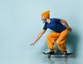 Elderly man in t-shirt, orange pants, hat, gumshoes. Riding black skateboard, posing sideways on blue background. Full length Royalty Free Stock Photo