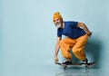 Elderly man in t-shirt, orange pants, hat, gumshoes. Riding black skateboard, posing sideways on blue background. Full length