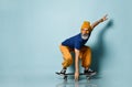 Bearded elderly man in t-shirt, sunglasses, orange pants, hat, gumshoes. Riding black skateboard, posing on blue background