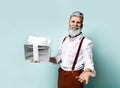 Bearded elderly man in white shirt, brown pants and suspenders. He is smiling, holding silver gift box, posing on blue background Royalty Free Stock Photo