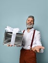 Bearded elderly man in white shirt, brown pants and suspenders. He is smiling, holding silver gift box, posing on blue background