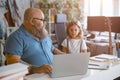 Bearded father looks at little girl taking part in video lesson via laptop at home