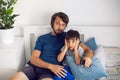 bearded father and son are sitting on the bed in T-shirts and watching football match on TV. a man holds a remote