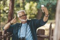 Bearded gray-haired European man takes a selfie photo on a smartphone in the park. The pensioner walks and enjoys life