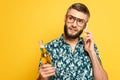 Bearded guy in glasses with refreshing bottle of beer talking on smartphone isolated on yellow