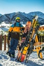 A bearded guy in a helmet and glasses stands near a rack with skis and snowboards. Beautiful sunny day at the ski resort