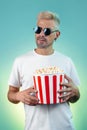 Bearded hipster blonde man wearing stylish sunglasses and holding bucket with popcorn in studio in neon lights. Royalty Free Stock Photo