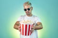 Bearded hipster blonde man wearing stylish sunglasses and holding bucket with popcorn in studio in neon lights. Royalty Free Stock Photo