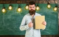 Bearded hipster holds book, chalkboard on background. Teacher in eyeglasses recommends book, thumbs up. Recommendation Royalty Free Stock Photo