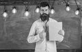 Bearded hipster holds book, chalkboard on background. Teacher in eyeglasses recommends book, thumbs up. Recommendation Royalty Free Stock Photo