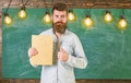 Bearded hipster holds book, chalkboard on background. Teacher in eyeglasses recommends book, thumbs up. Recommendation Royalty Free Stock Photo