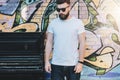 Bearded hipster man dressed in white t-shirt is stands against wall with graffiti. Mock up. Space for logo, text, image.