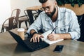 Bearded hipster man sits at table in front of laptop and writes in notebook,student prepares for exams,freelancer works. Royalty Free Stock Photo