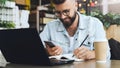 Bearded hipster man sits at table in front of laptop and writes in notebook,student prepares for exams,freelancer works. Royalty Free Stock Photo