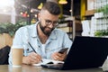 Bearded hipster man sits at table in front of laptop and writes in notebook,student prepares for exams,freelancer works. Royalty Free Stock Photo