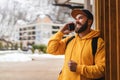 Bearded hipster man tourist in yellow hoodie and cap stands outdoors, talking on mobile phone. Smiling man calling friends