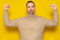 Bearded Hispanic man wearing a beige turtleneck showing his biceps isolated over yellow background, he is in lousy shape