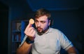 Bearded hungry man sits at night in a room with a blue light on his desk and looks at the chips in his hand with a serious face.