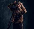 Bearded hunter with rifle and binoculars corrects his hat and looking sideways. Studio photo against a dark wall