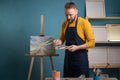 A bearded male artist dressed in a black apron stands in an art studio holding a palette with paints in his hands looks Royalty Free Stock Photo