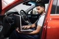 Bearded male mechanic sitting inside car using laptop, recording automobile engine checks