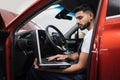 Bearded male mechanic sitting inside car using laptop, recording automobile engine checks