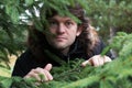 Bearded man with big round goggle eyes looking out of the forest through the young branches of fir trees. Scared man in