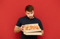 Bearded man in a black T-shirt is holding a box of fresh pizza and staring at a fast food on a red background with a hungry look. Royalty Free Stock Photo