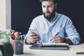 Bearded man doing work at desk. Male hipster holding smartphone and pen in his hands. Businessman taking notes, messaging on phone Royalty Free Stock Photo