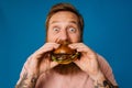 Bearded man eating burger while standing isolated over blue background