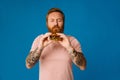 Bearded man eating burger while standing isolated over blue background