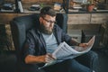 Bearded man in eyeglasses reading newspaper while sitting in armchair at home Royalty Free Stock Photo