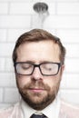 Bearded man in glasses, shirt and tie stands in bathroom under the warm water that comes from the watering can and with droplets