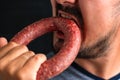 A bearded man greedily bites a round sausage. Hunger and gluttony