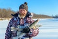 Bearded man is holding frozen fish after successful winter fishing at cold sunny day