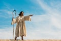 Man holding wooden cane and pointing with finger in desert against sky