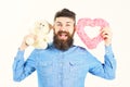 Bearded man holds symbols of love. Hipster with heart, teddy bear and happy joyful face. Macho with beard smiles Royalty Free Stock Photo