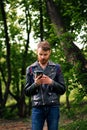 A bearded man looks thoughtfully at the phone while standing in a forest in blue waits and a black leather jacket